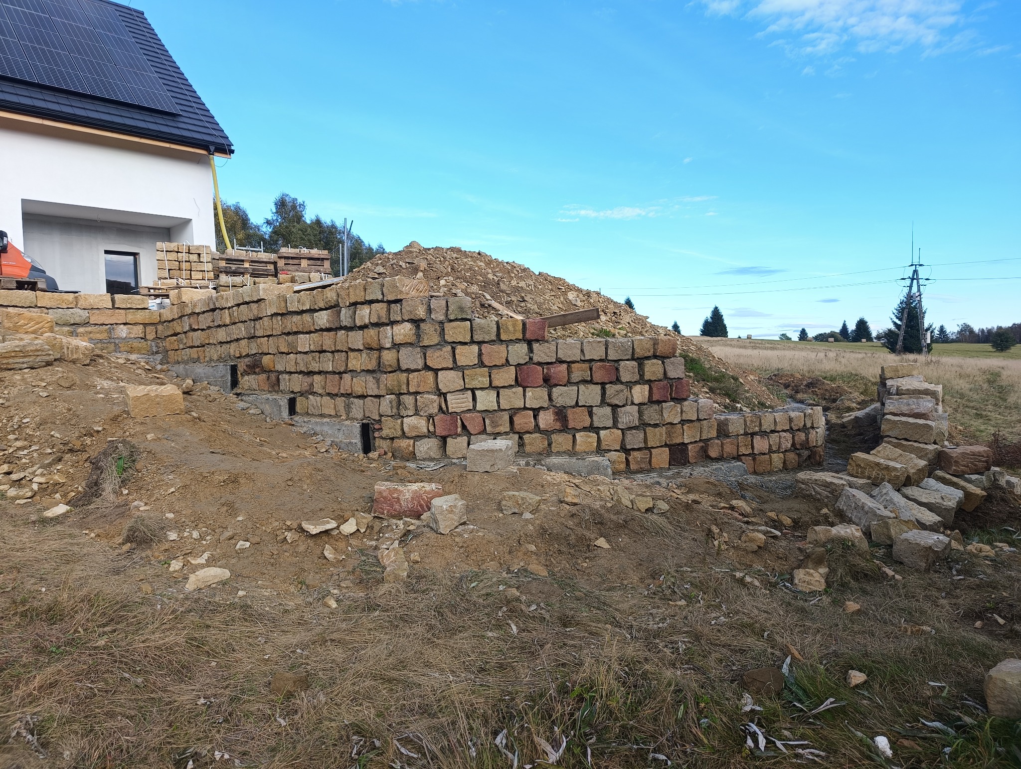 Wznoszenie muru oporowego z kamienia naturalnego przy nowym domu z panelami słonecznymi na dachu; widoczne fundamenty z otworami wentylacyjnymi.