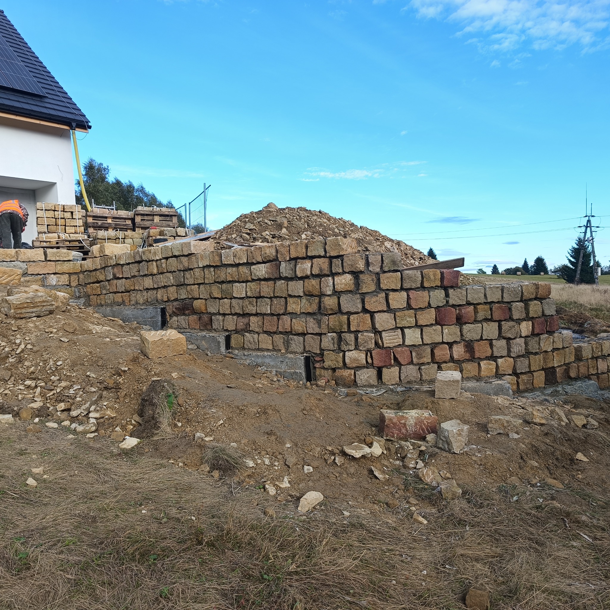 Wznoszenie muru oporowego z kamiennych bloków przy nowo budowanym domu z panelami słonecznymi na dachu, widoczne fundamenty i stos kamieni na ziemi.