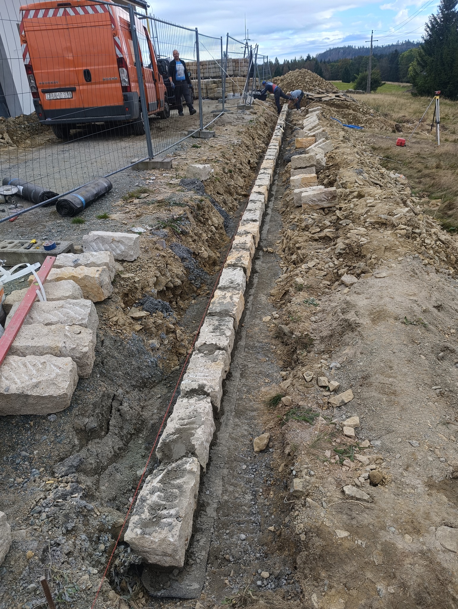 Wykop pod fundament z ułożonymi blokami kamiennymi, wyznaczony sznurkiem, na tle budowy z pomarańczowym busem i pracującymi osobami.