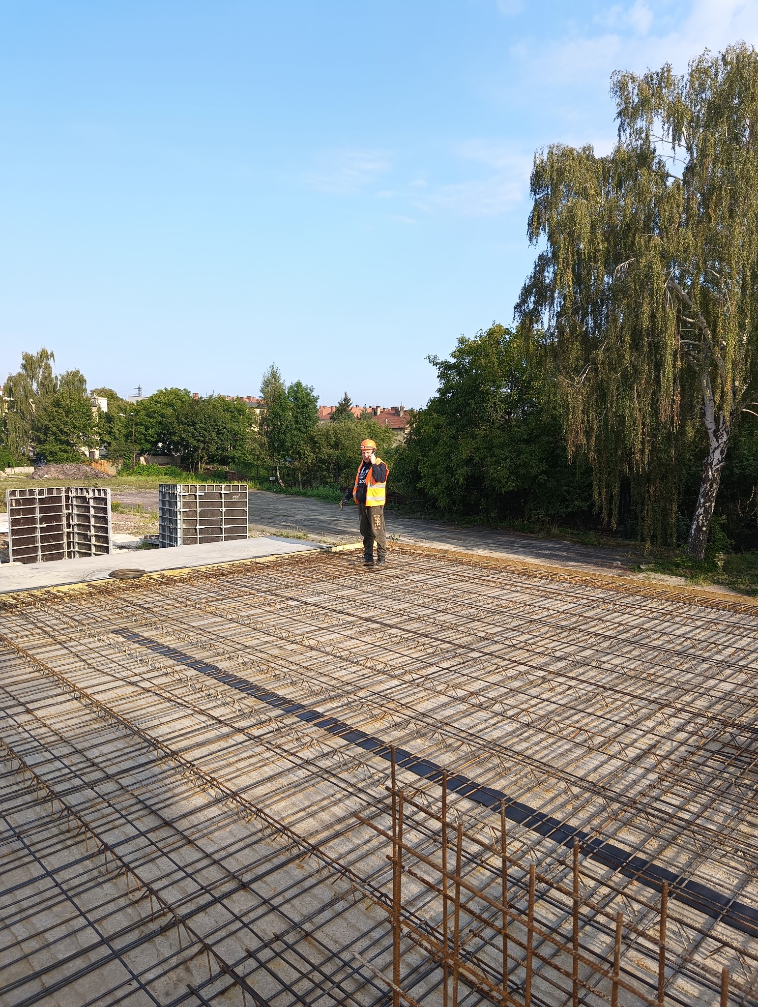 Zbrojenie fundamentu domu jednorodzinnego z widocznym pracownikiem w pomarańczowej kamizelce i kasku, stojącym na zbrojeniu i rozmawiającym przez telefon, w tle drzewa i budynki mieszkalne.