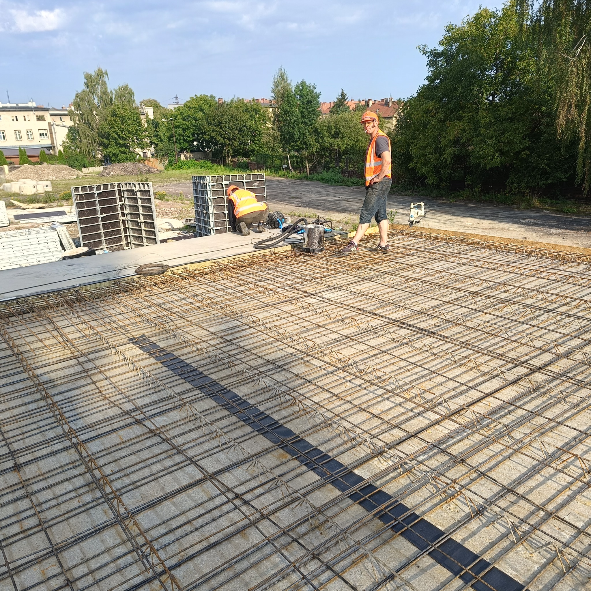 Zbrojenie płyty fundamentowej na placu budowy z pracownikami w odblaskowych kamizelkach i pomarańczowych kaskach, widoczne szalunki i narzędzia.