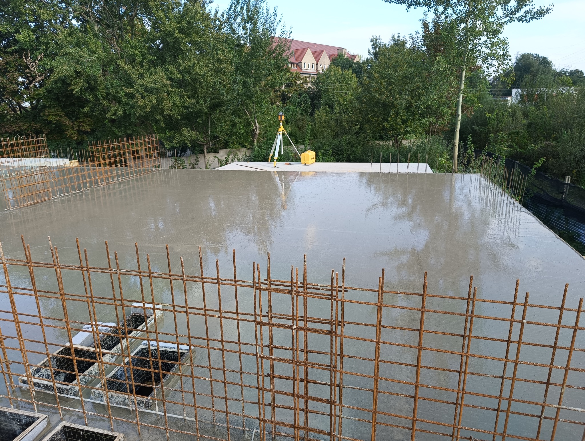 Świeżo wylany, mokry beton na płycie fundamentowej z widocznym zbrojeniem i niwelatorem laserowym w tle.
