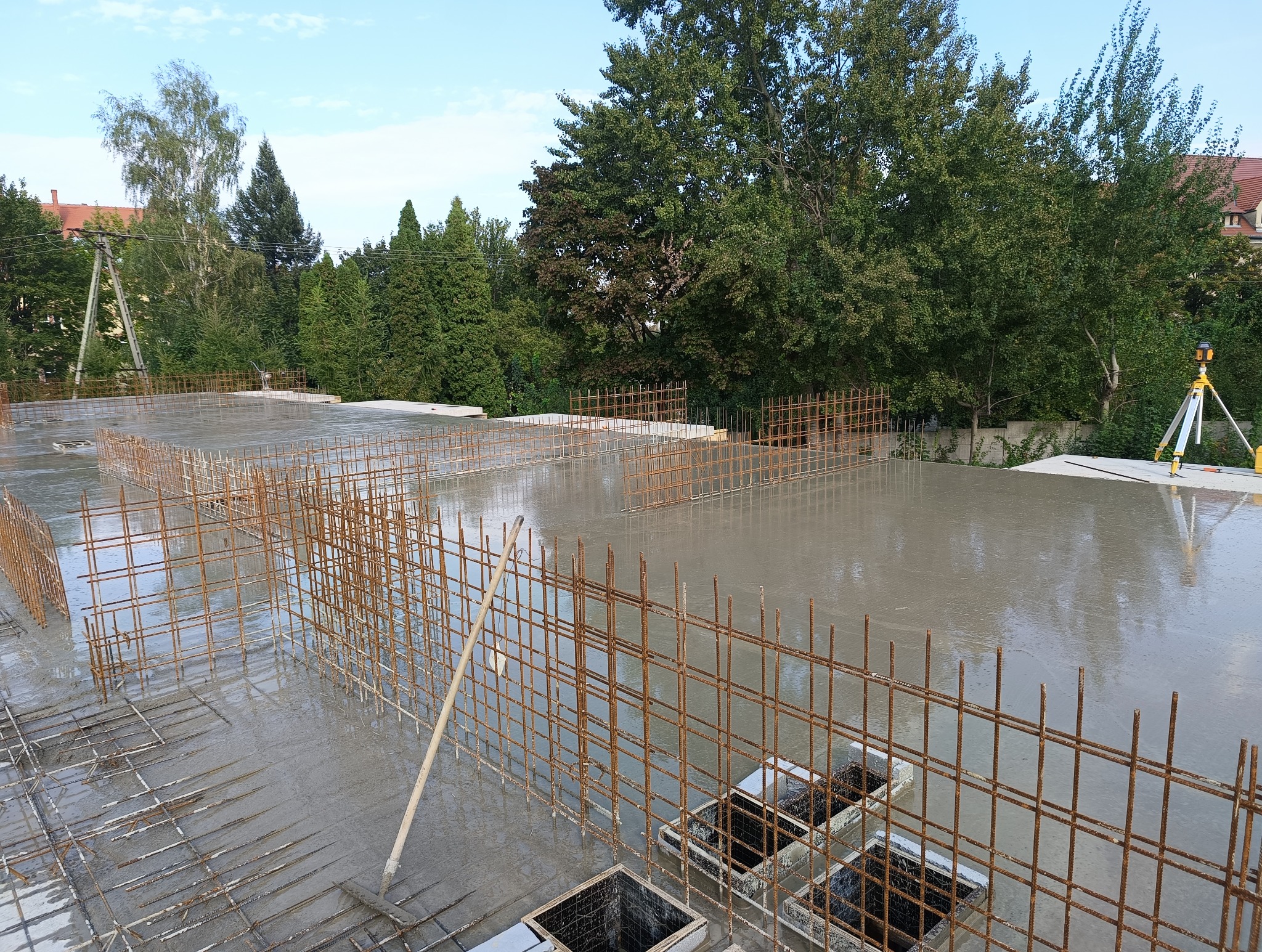 Zalany betonem fundament z wystającymi prętami zbrojeniowymi, widoczny niwelator laserowy na trójnogu i otaczająca zieleń.