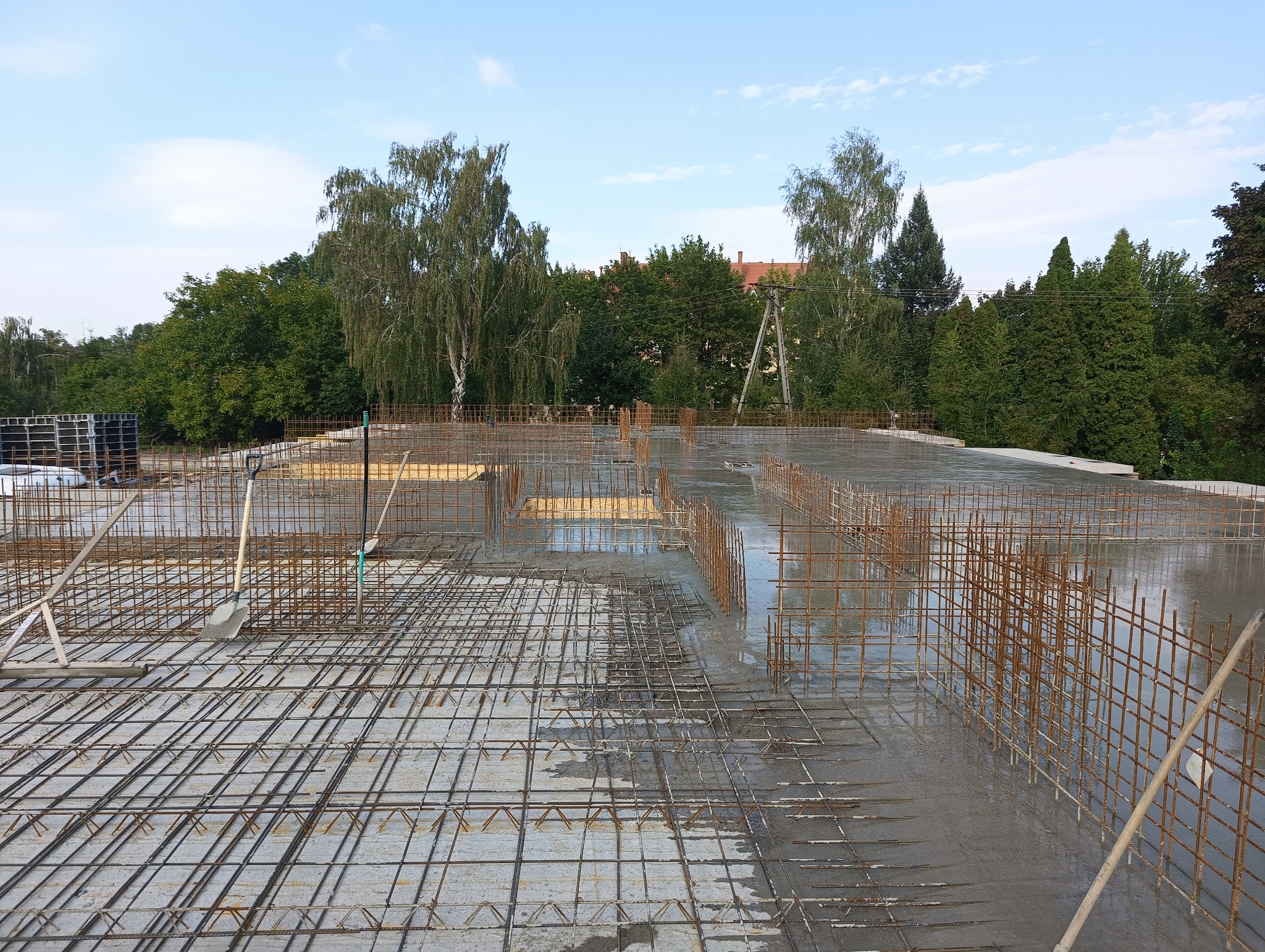 Zalany betonem strop z widocznym zbrojeniem, szpadel i grabie na powierzchni betonu, w tle zieleń drzew i budynki.