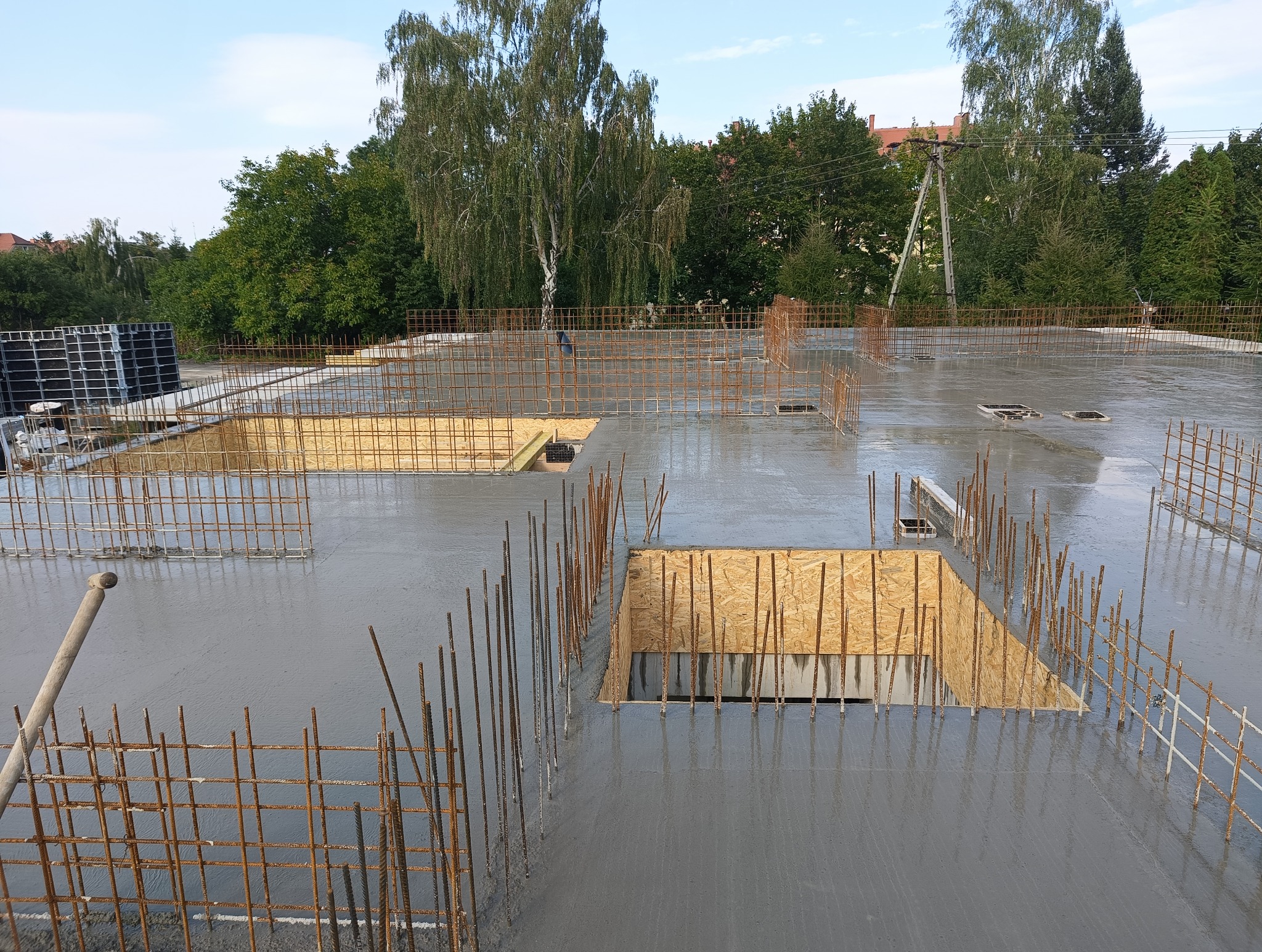 Świeżo wylany, mokry betonowy strop z wystającymi prętami zbrojeniowymi i drewnianymi szalunkami otworów, widok z góry na tle zieleni.