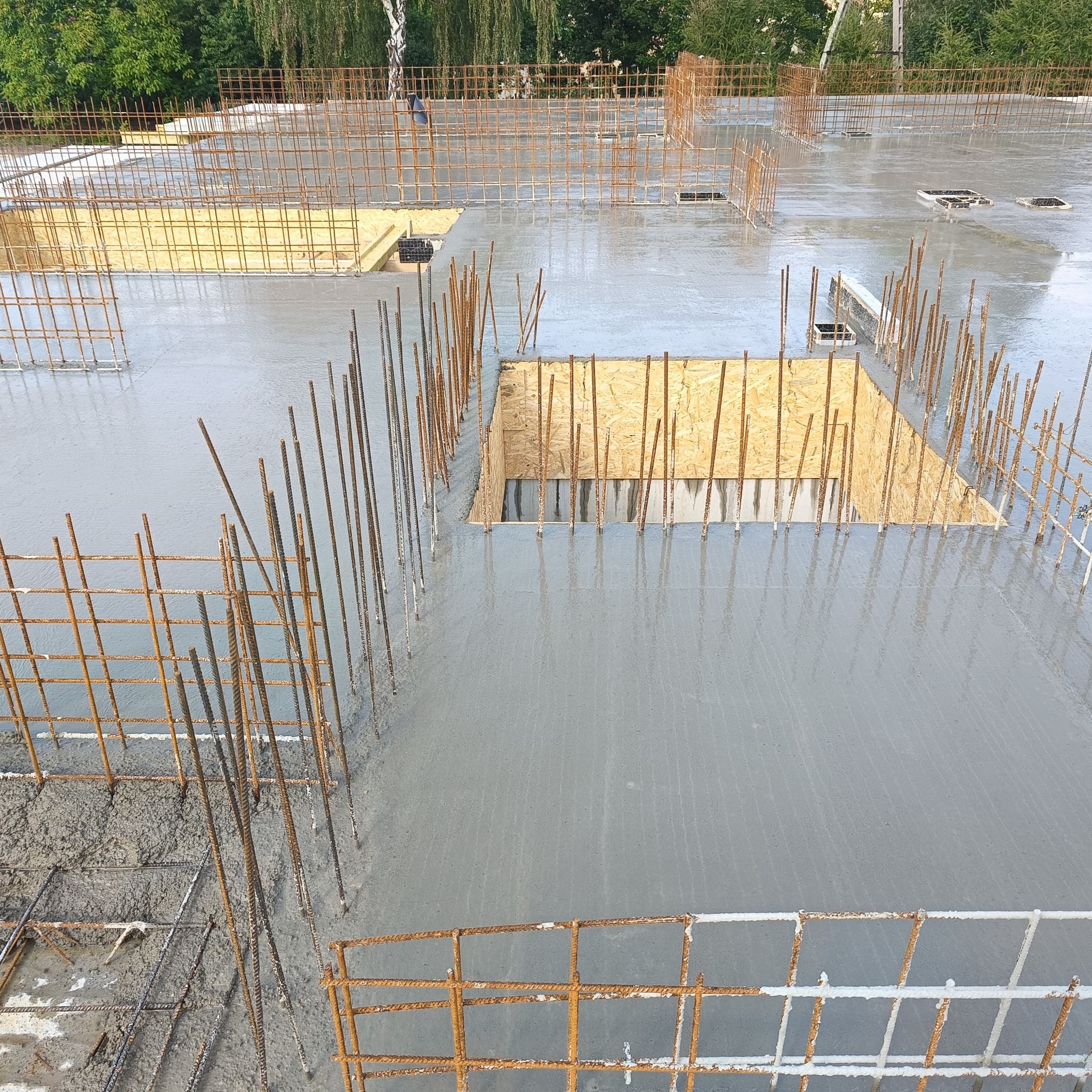 Świeżo wylany beton na stropie z widocznym zbrojeniem i otworami szalunkowymi wyłożonymi sklejką; w tle zieleń drzew.