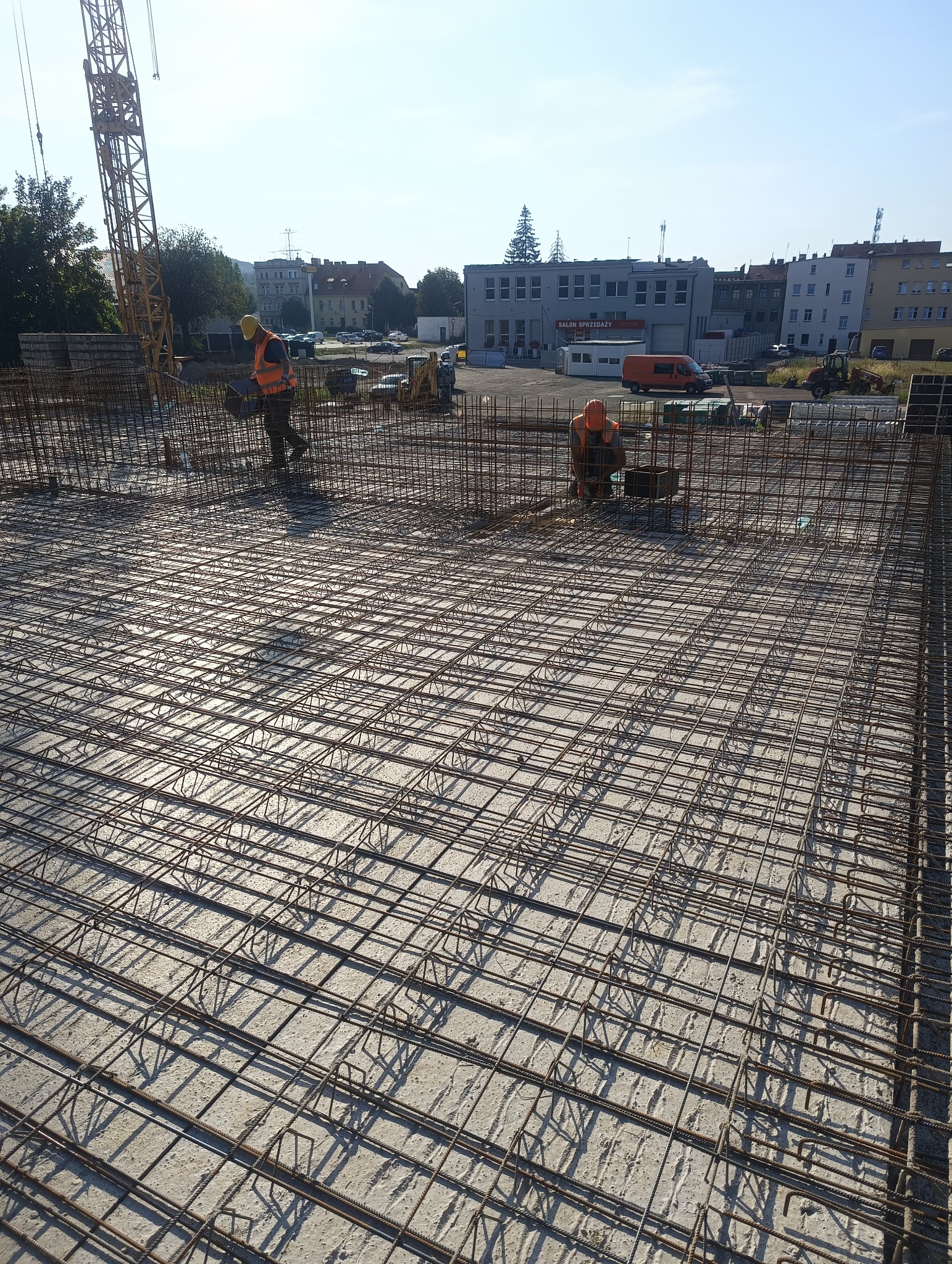 Siatka zbrojeniowa na betonowym fundamencie, dwóch robotników w pomarańczowych kamizelkach montuje zbrojenie, w tle budynki miejskie i żuraw.