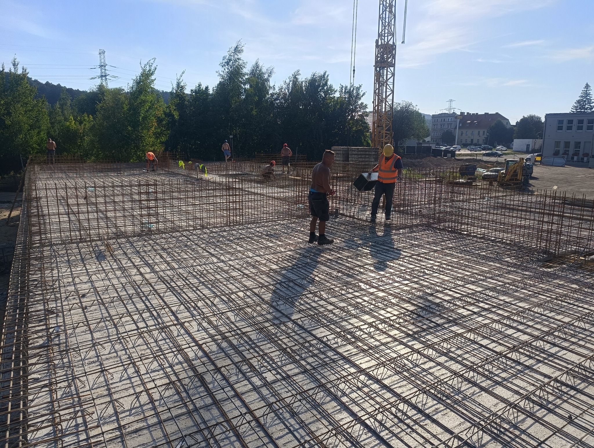 Zbrojenie fundamentu płyty betonowej z widocznymi pracownikami w kamizelkach odblaskowych i kasku, w tle budynki mieszkalne i dźwig budowlany.
