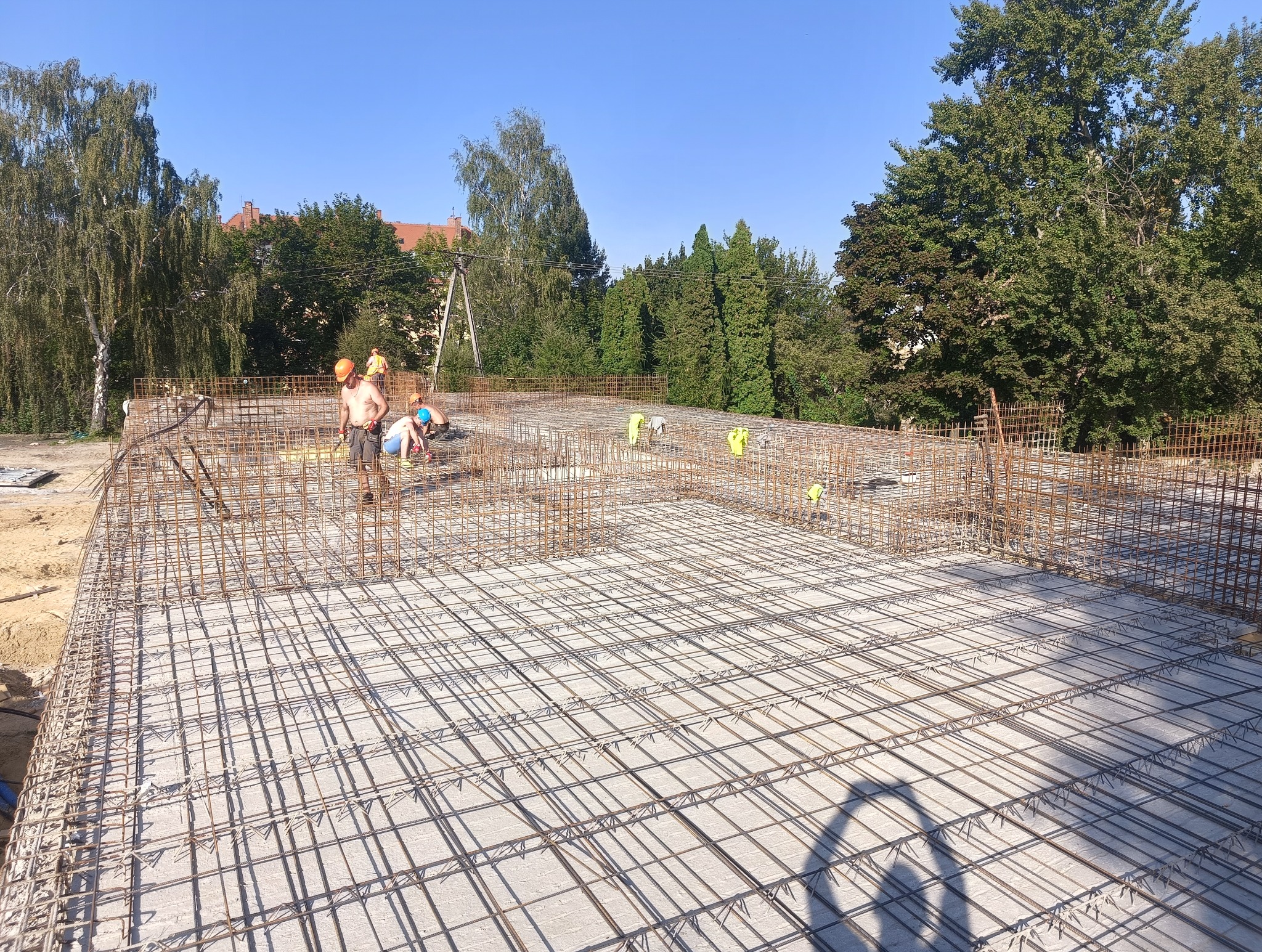 Zbrojenie płyty fundamentowej z widocznymi pracownikami w kaskach i kamizelkach odblaskowych, przygotowane do zalania betonem, w tle zieleń drzew i budynki.