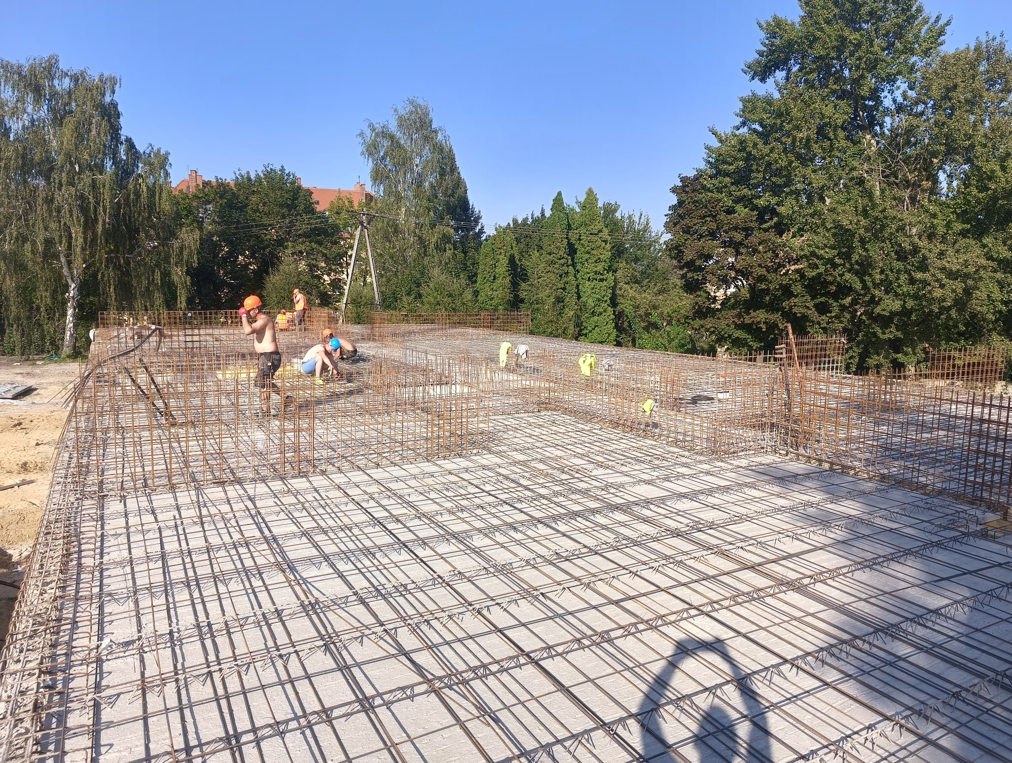 Zbrojenie fundamentów na budowie domu w Nowym Julianowie, z pracownikami w kaskach i bez koszulek, widoczne drzewa w tle.
