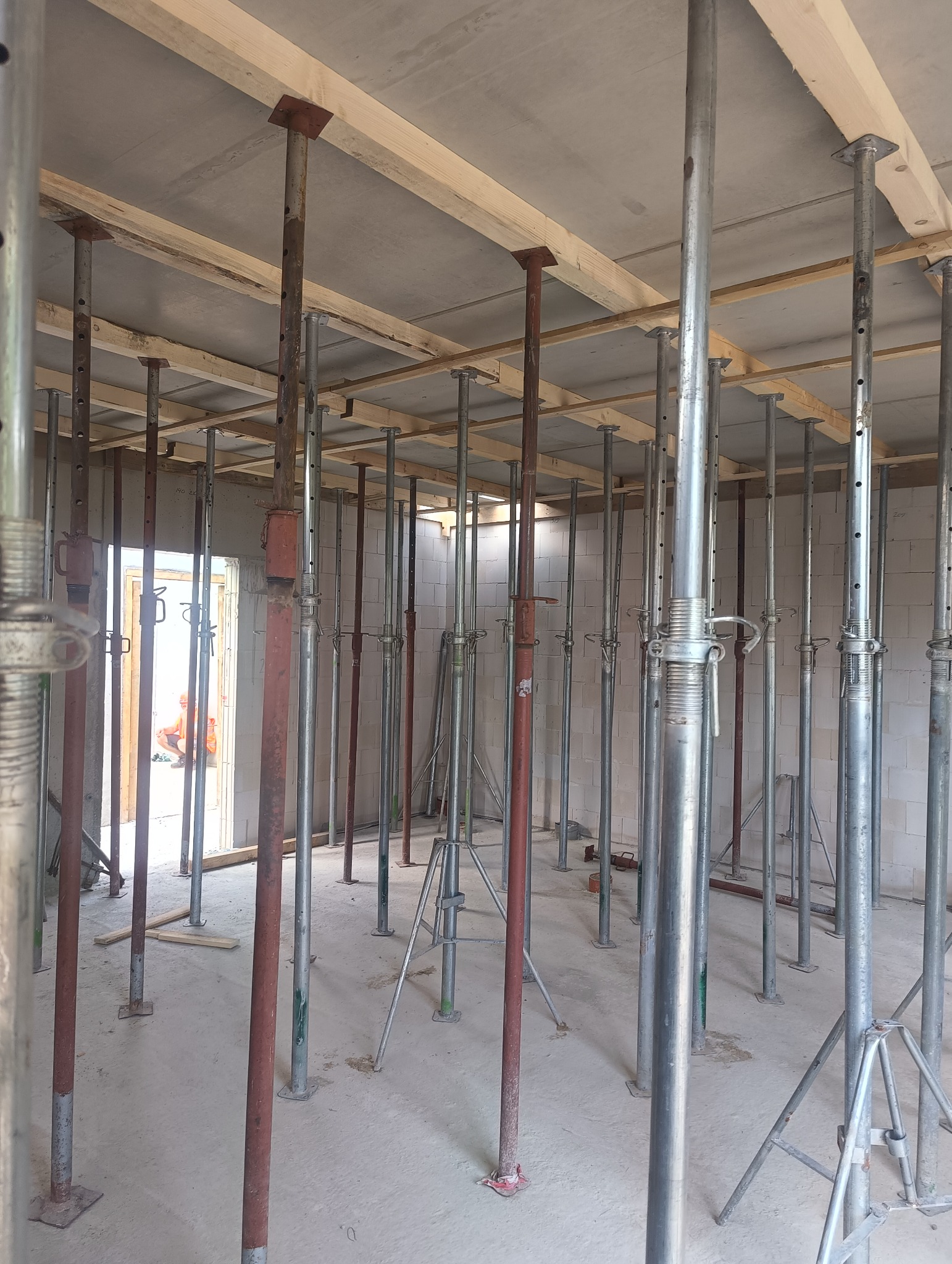 Wnętrze w trakcie budowy z betonowym sufitem podpartym drewnianymi belkami i metalowymi stemplami budowlanymi, widoczny mur z białych bloczków.