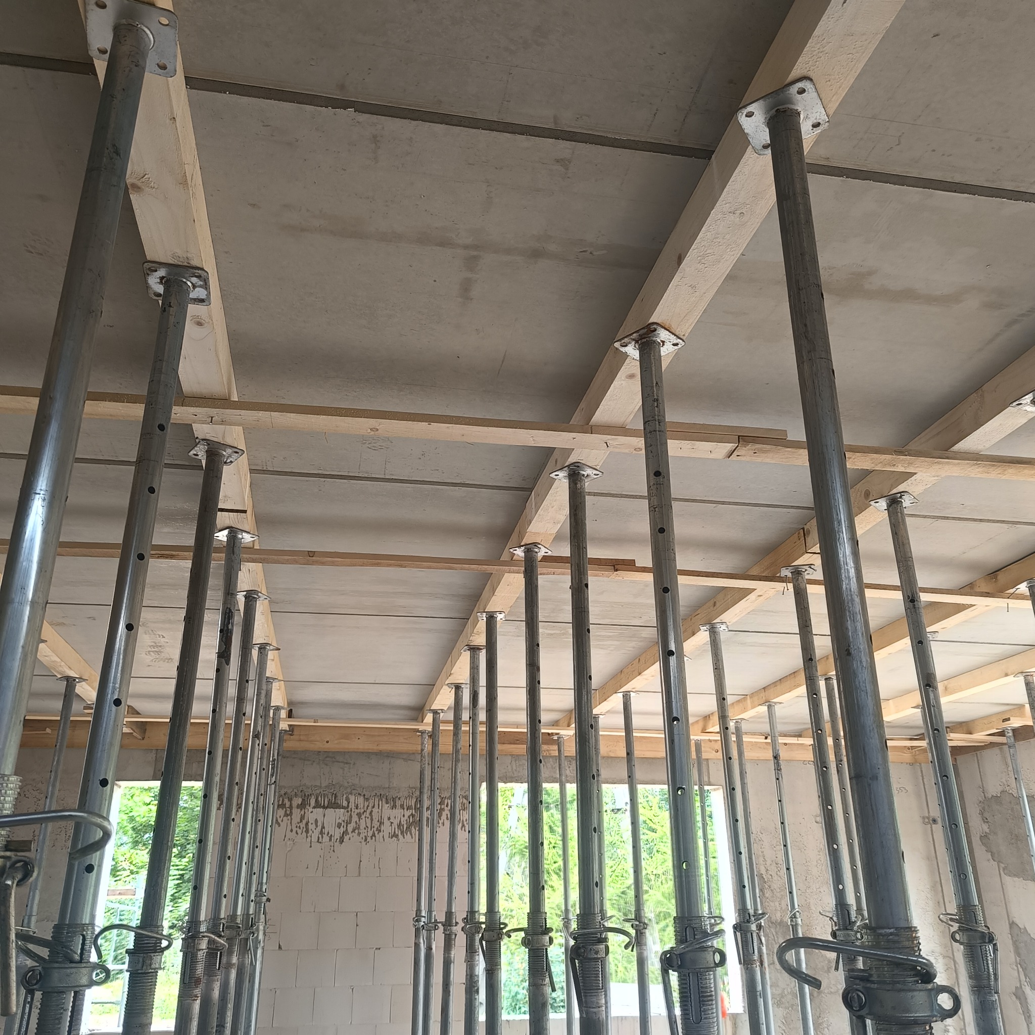 Szare metalowe stemple budowlane podtrzymujące drewniane belki stropowe na placu budowy, widok z dołu.