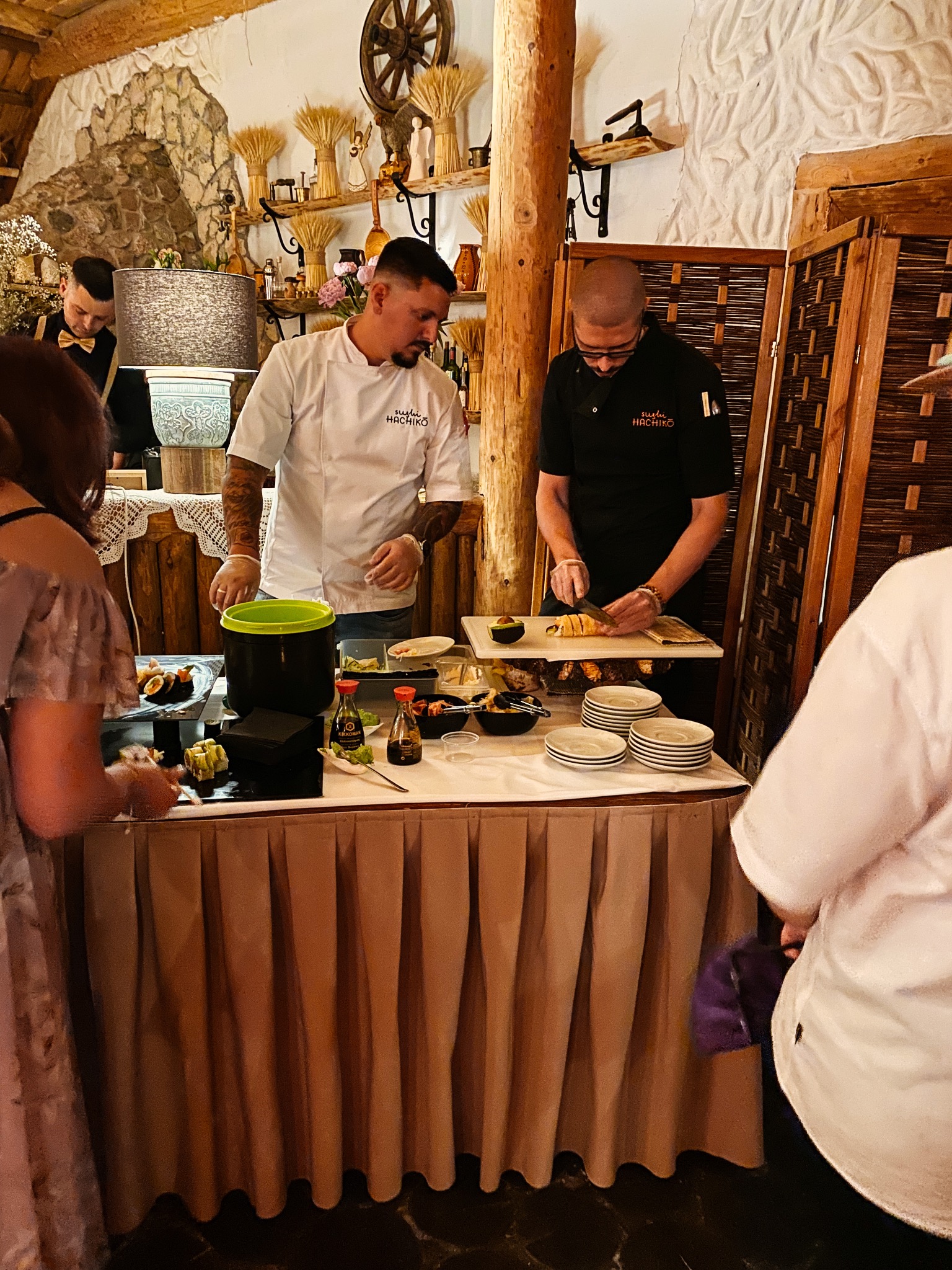 Dwóch kucharzy w firmowych uniformach przygotowuje sushi na stole podczas wydarzenia, z widocznymi gośćmi i elementami dekoracyjnymi w tle.