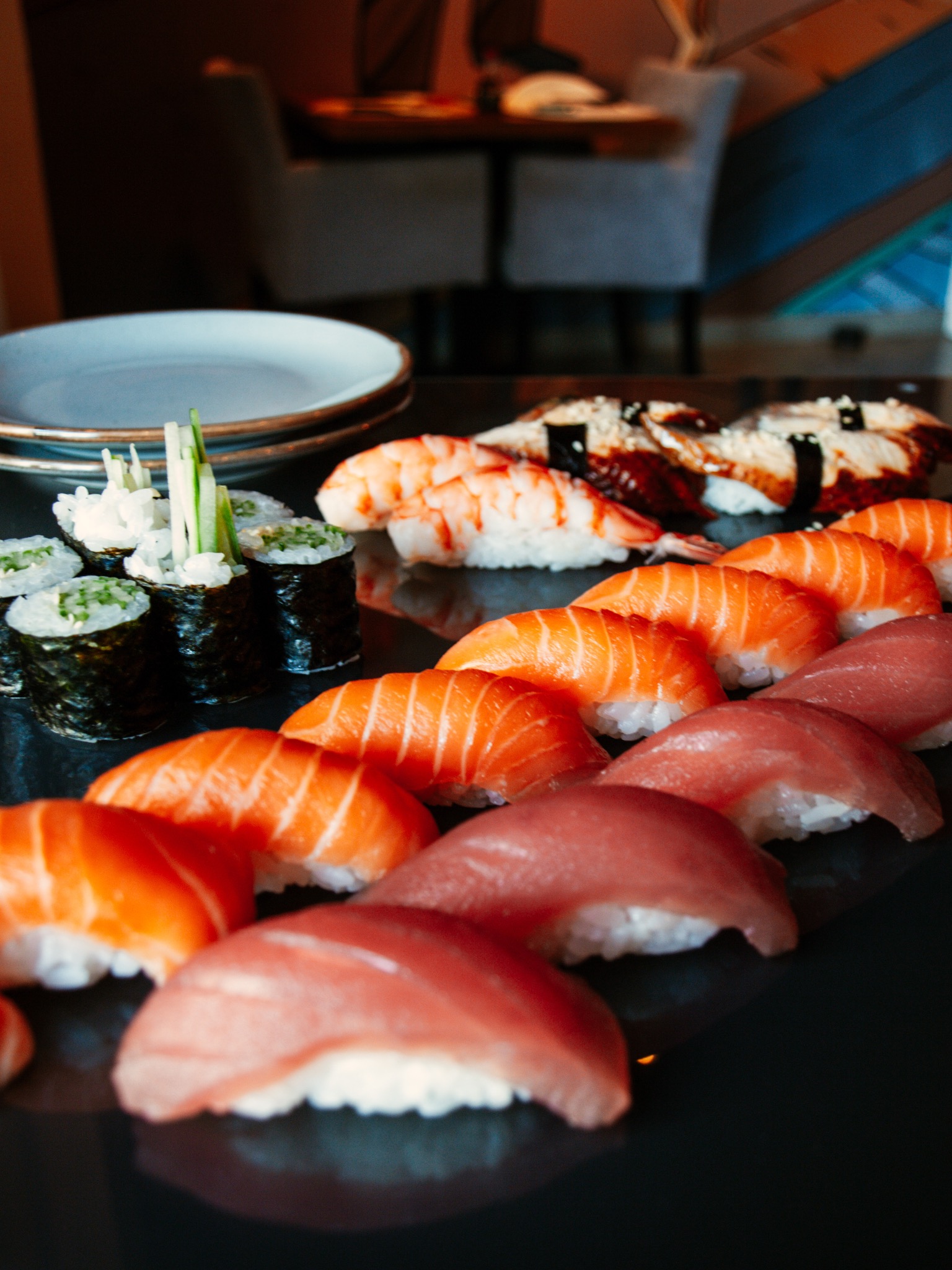 Zestaw sushi z tuńczykiem, łososiem, krewetką i węgorzem, ułożony na czarnym, błyszczącym talerzu, z dodatkiem maki z ogórkiem i rzodkwią, talerze w tle.