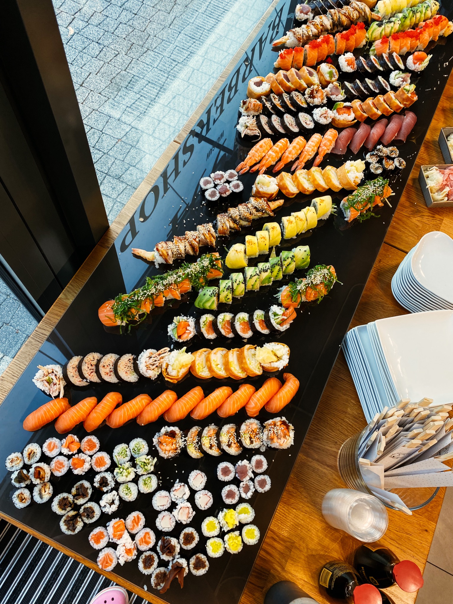Bogaty wybór sushi i rolek ułożonych na czarnym, błyszczącym stole obok okna, z widocznymi naczyniami, sosami i pałeczkami.