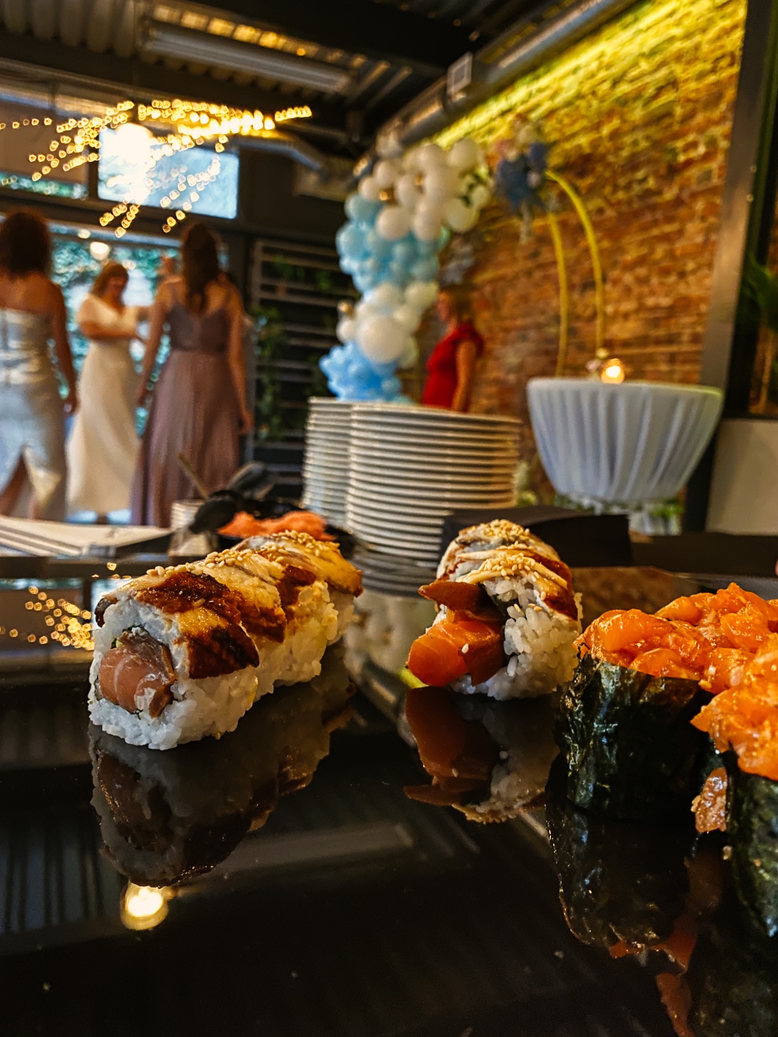Sushi z łososiem i węgorzem na czarnym, błyszczącym stole, w tle dekoracja balonowa i goście na przyjęciu.