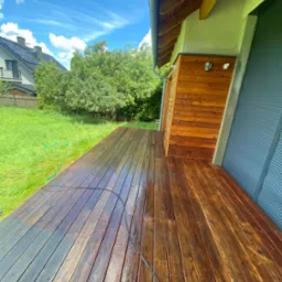 Impregnowana drewniana deska tarasowa, częściowo mokra, z widocznym wężem ogrodowym i fragmentem elewacji budynku z drewnianą boazerią, w tle trawnik i dom jednorodzinny