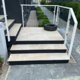 Wyłożone jasnymi płytkami schody zewnętrzne z białą balustradą i szybami, na górnym stopniu leży opona, obok schodów skrzynka elektryczna i rower.