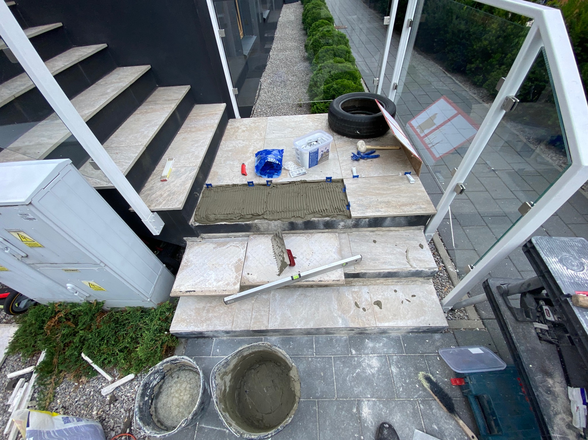 Montaż płytek na schodach zewnętrznych, widoczna warstwa kleju, poziomica, narzędzia glazurnicze i wiadro z zaprawą, w tle zielone krzewy i fragment elewacji budynku.