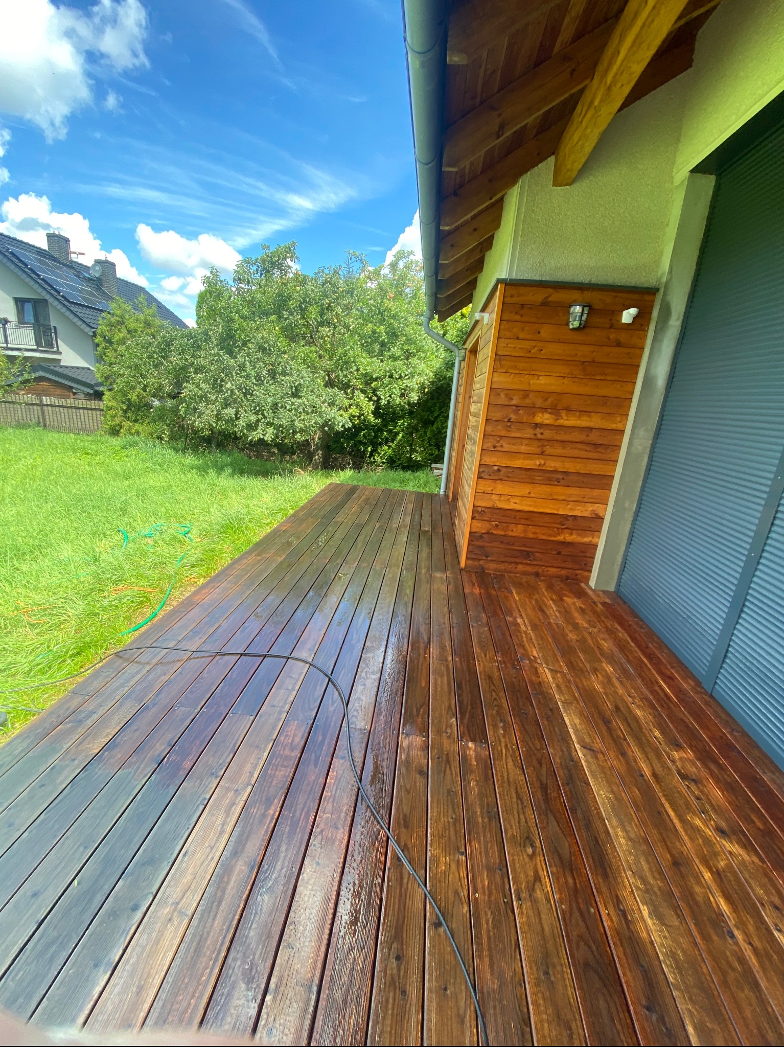 Impregnowana drewniana deska tarasowa, częściowo mokra, z widocznym wężem ogrodowym i fragmentem elewacji budynku z drewnianą boazerią, w tle trawnik i dom jednorodzinny