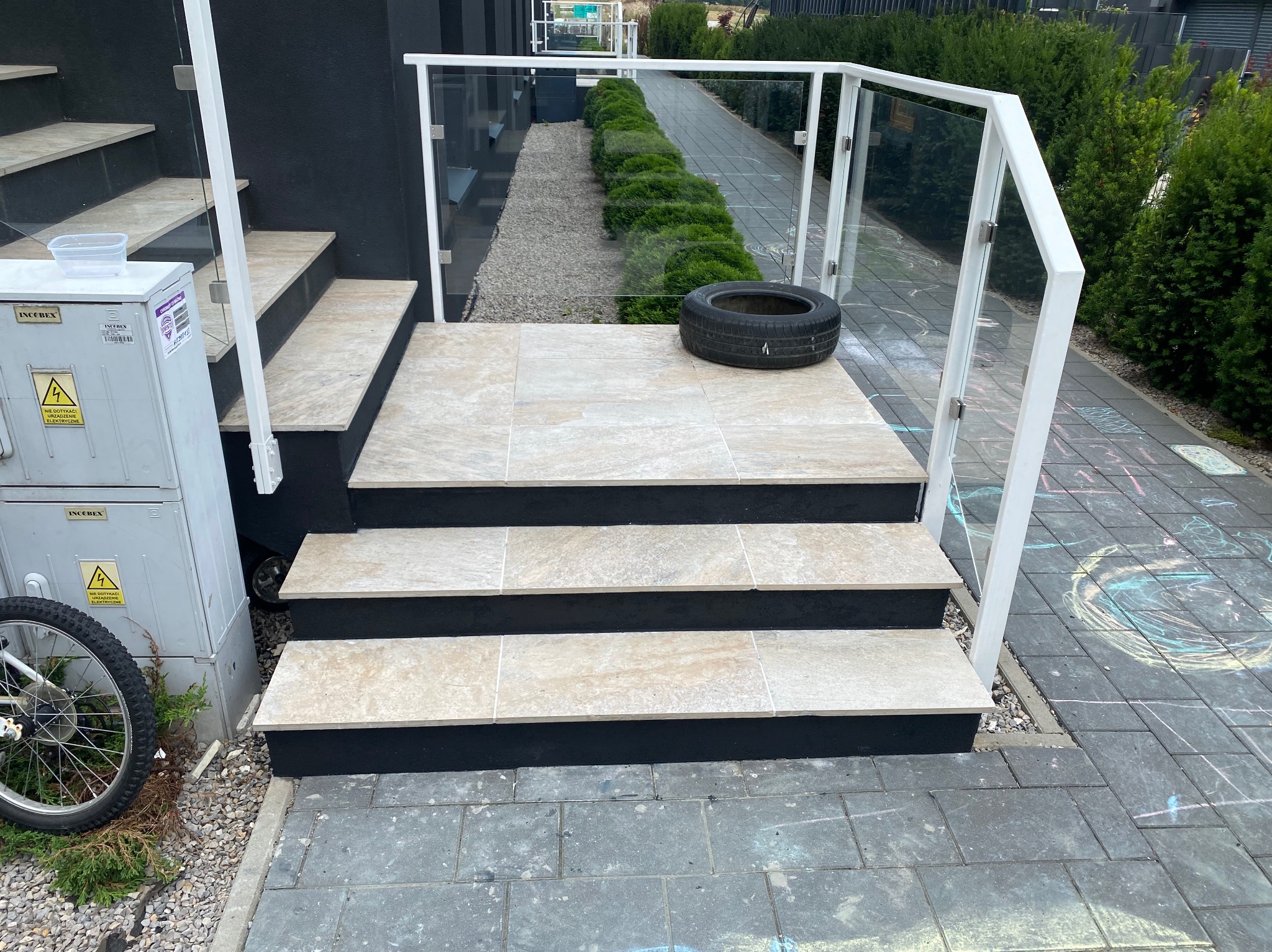 Wyłożone jasnymi płytkami schody zewnętrzne z białą balustradą i szybami, na górnym stopniu leży opona, obok schodów skrzynka elektryczna i rower.