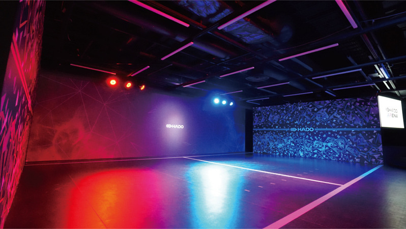 Wnętrze hali sportowej oświetlone neonowymi światłami w odcieniach różu, fioletu i niebieskiego, z logo HADO na ścianie i liniami wyznaczającymi pole gry.