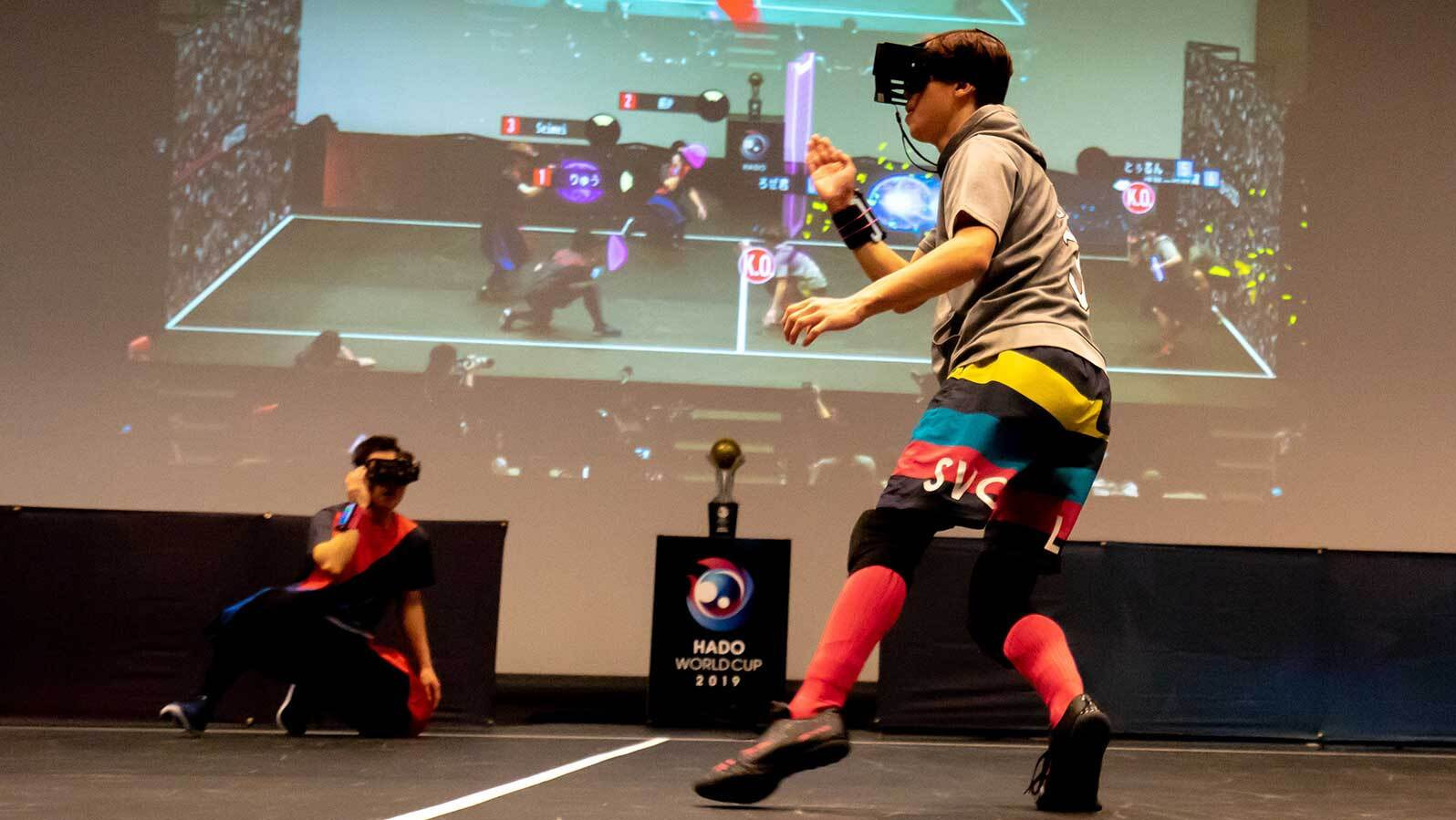 Mężczyzna w okularach VR gra w wirtualną grę zespołową na scenie podczas turnieju, w tle wyświetlany jest obraz z gry i logo HADO World Cup 2019.
