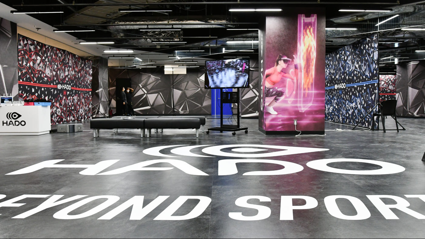 Przestronne wnętrze hali HADO z grafikami na ścianach, napisem 'HADO BEYOND SPORT' na podłodze, ławkami i ekranem prezentującym rozgrywkę VR.