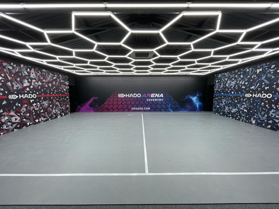 Wnętrze areny HADO z geometrycznym oświetleniem sufitowym i ścianami w kolorach czerwonym i niebieskim, z logo i nazwą areny na ścianie w tle.