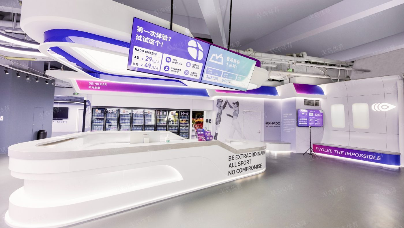 Nowoczesne wnętrze centrum rozrywki sportowej z ladą recepcyjną, ekranami informacyjnymi z cenami i ofertami, chłodziarkami z napojami oraz grafiką ścienną przedstawiającą osoby w okularach VR.