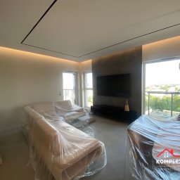 KompleXoweWykończenia - Salon w trakcie zabezpieczania mebli folią ochronną przed pracami wykończeniowymi, widoczne sofy i stół przykryte folią, w tle okno z widokiem na zieleń miejską i nowoczesne oświetlenie sufitowe.