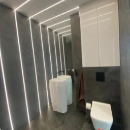 Nowoczesna łazienka z szarymi płytkami, podświetleniem LED wzdłuż ścian i sufitu, białą umywalką wolnostojącą, wiszącą toaletą i szafką nad nią.