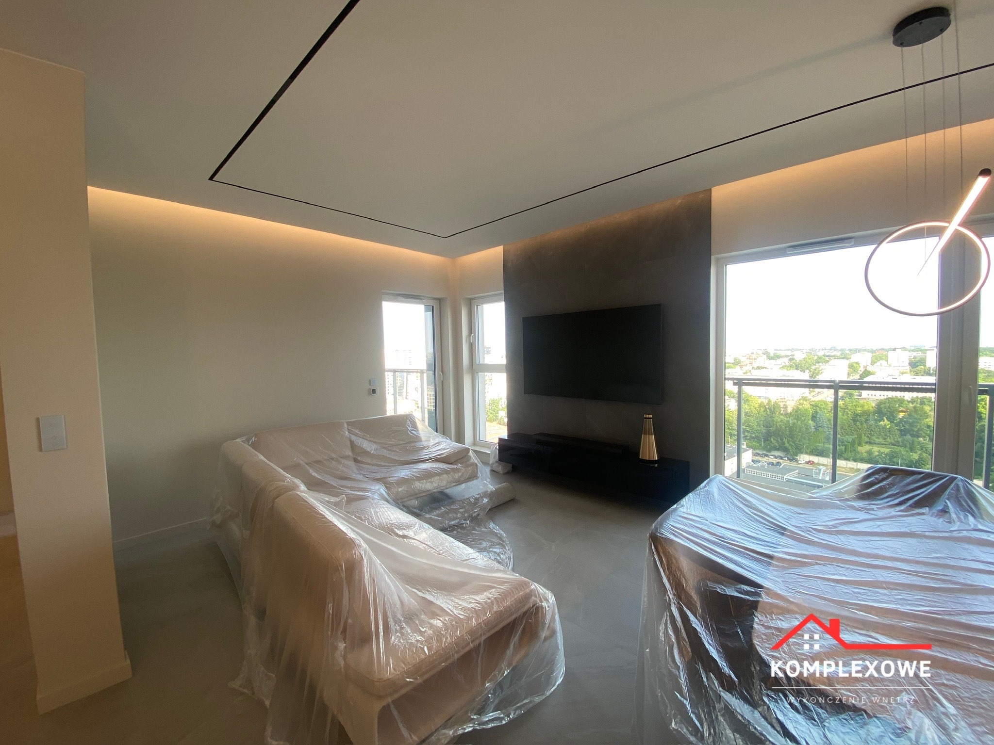 Salon w trakcie zabezpieczania mebli folią ochronną przed pracami wykończeniowymi, widoczne sofy i stół przykryte folią, w tle okno z widokiem na zieleń miejską i nowoczesne oświetlenie sufitowe.