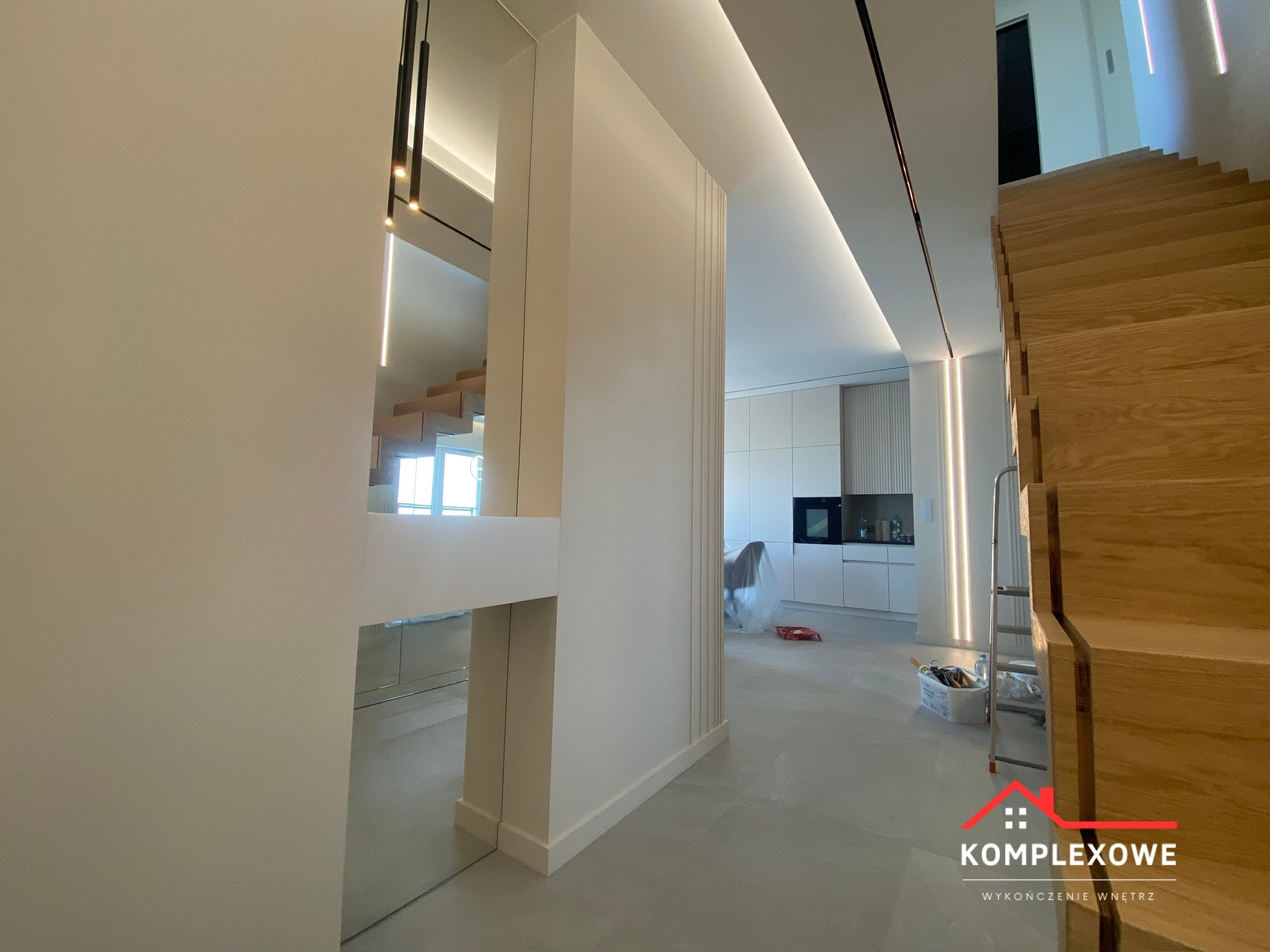 Nowoczesne wnętrze domu w trakcie wykończenia: drewniane schody, białe ściany z subtelnymi dekoracjami, minimalistyczna kuchnia w tle, widoczne zabezpieczenia folią malarską i drabina.