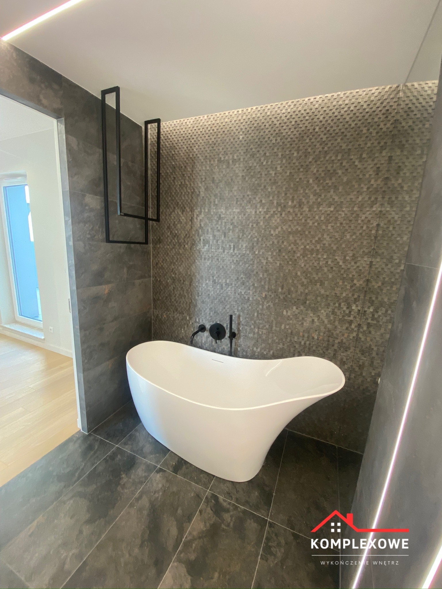 Nowoczesna łazienka z wolnostojącą białą wanną o asymetrycznym kształcie, czarna armatura, ściana wyłożona mozaiką w odcieniach szarości, podświetlenie LED i dekoracyjny element w formie czarnych...