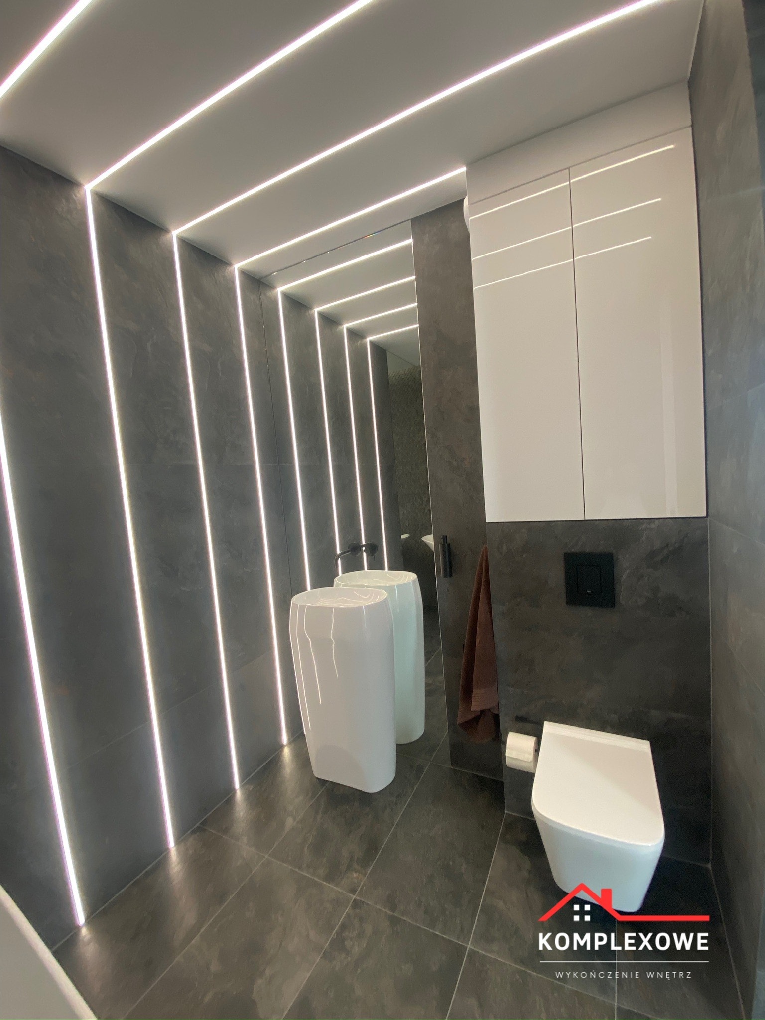 Nowoczesna łazienka z szarymi płytkami, podświetleniem LED wzdłuż ścian i sufitu, białą umywalką wolnostojącą, wiszącą toaletą i szafką nad nią.