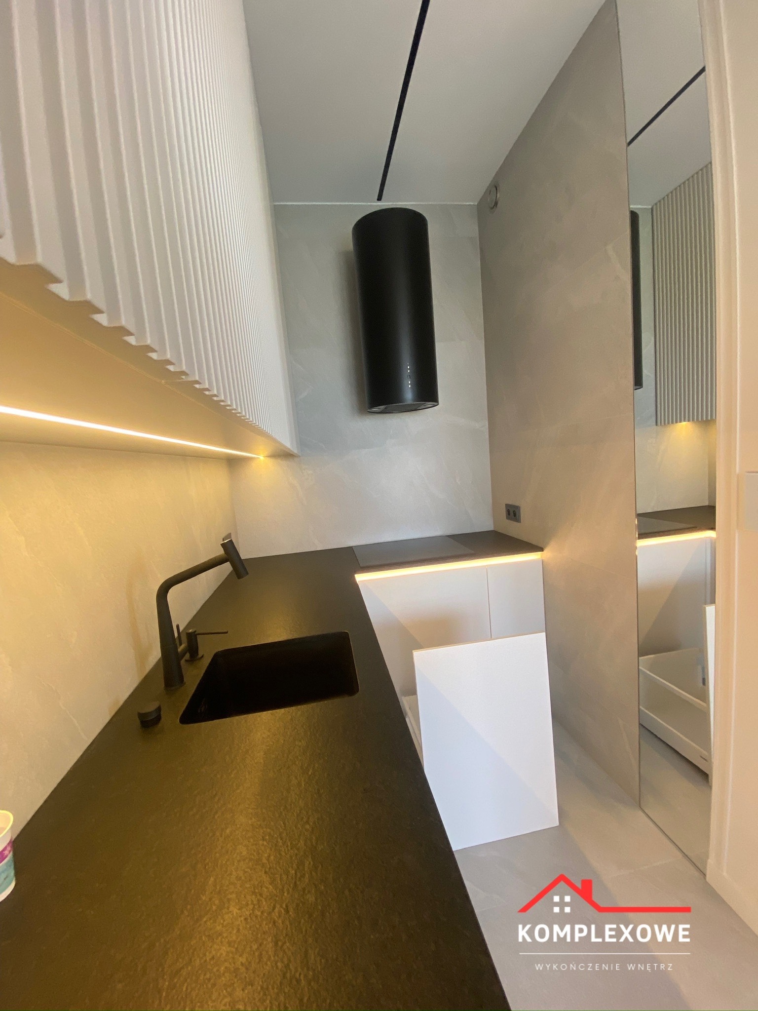 Nowoczesna kuchnia z czarnym blatem, czarną armaturą i okapem cylindrycznym, podświetleniem LED i szarymi płytkami imitującymi kamień, odbicie w lustrzanej ścianie