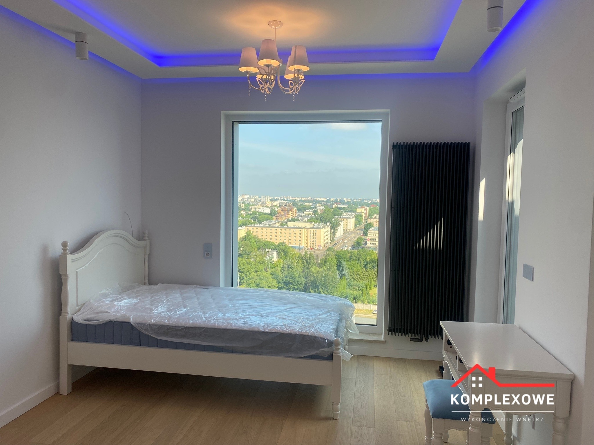 Jasne wnętrze pokoju z białymi ścianami, drewnianą podłogą i dużym oknem z widokiem na miasto, z białym łóżkiem z materacem owiniętym folią, czarnym grzejnikiem i białą toaletką z niebieskim pufem,...