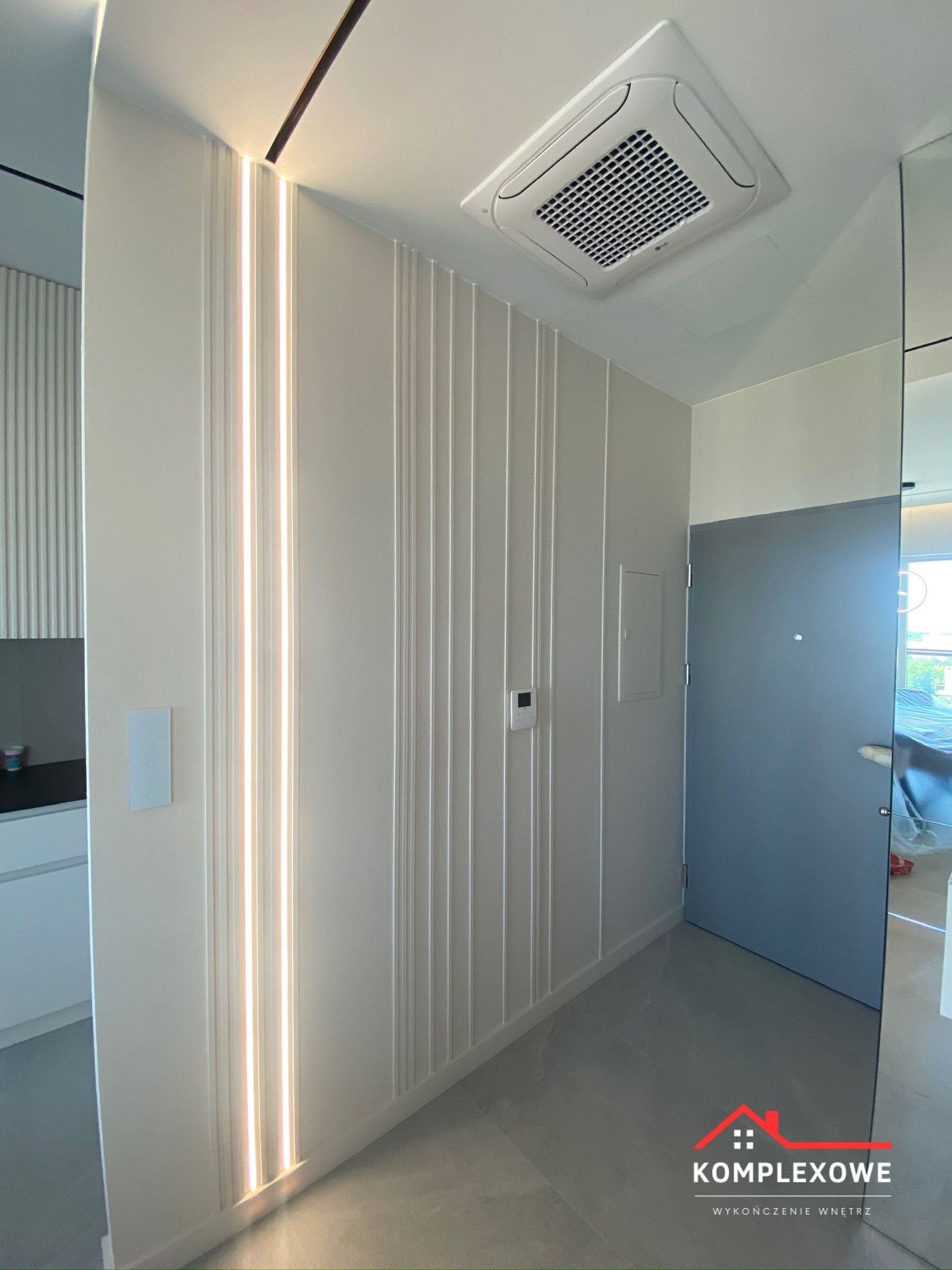 Nowoczesny przedpokój z dekoracyjną ścianą z pionowymi panelami i oświetleniem LED, szara podłoga, klimatyzator kasetonowy na suficie, widoczne drzwi wejściowe i fragment wnętrza mieszkania.