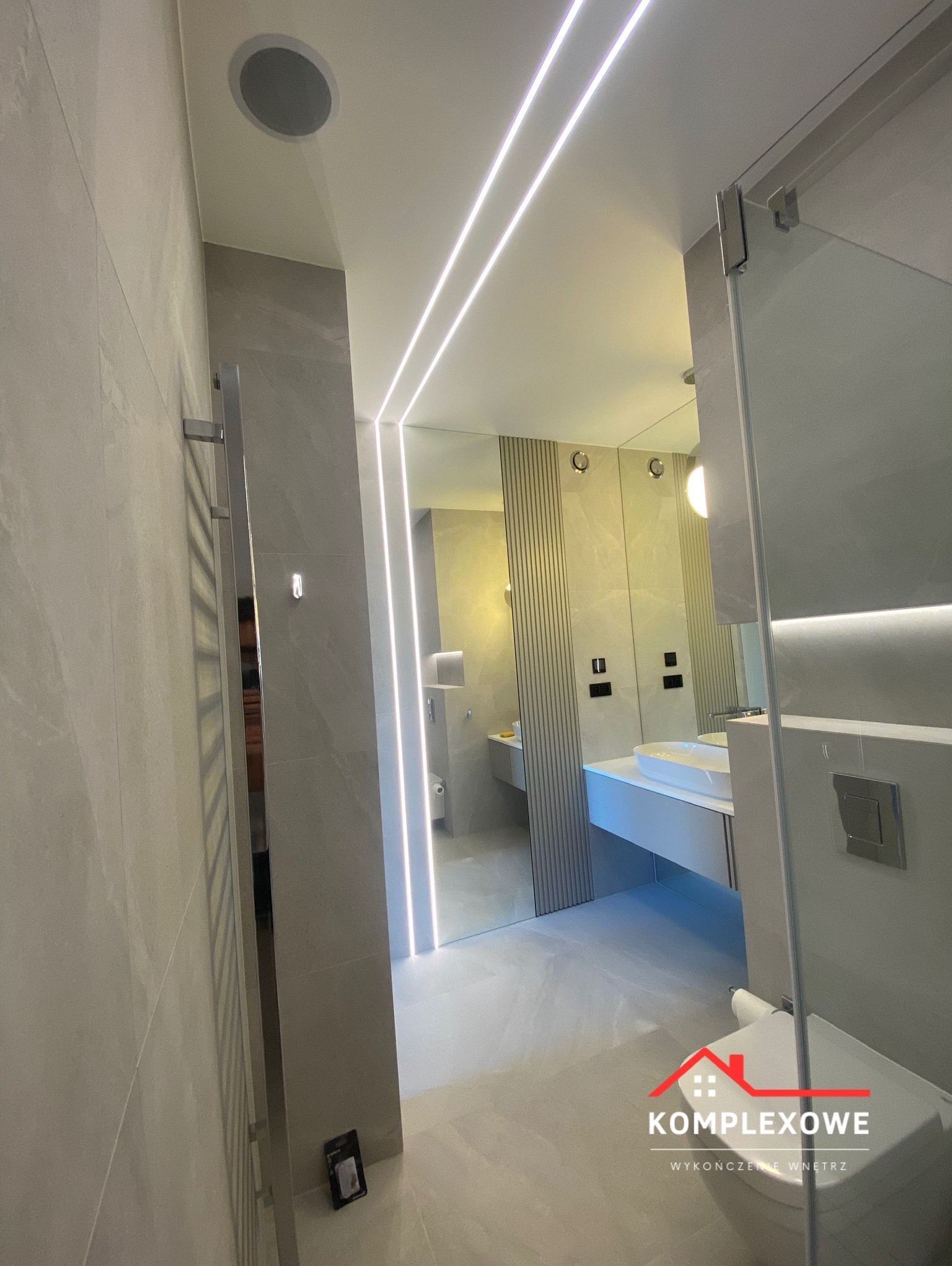 Nowoczesna łazienka w odcieniach szarości z podświetleniem LED na suficie, widoczna kabina prysznicowa z przezroczystego szkła, grzejnik drabinkowy na ścianie i logo firmy w dolnym rogu.