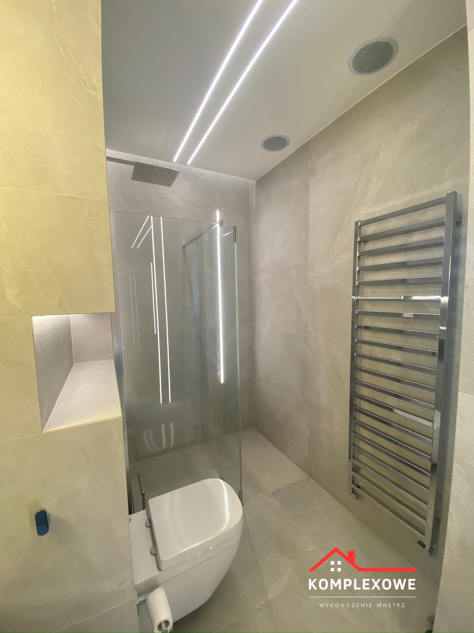 Nowoczesna łazienka z kabiną prysznicową typu walk-in, toaletą podwieszaną, grzejnikiem drabinkowym i oświetleniem LED w suficie podwieszanym, wykończona płytkami w odcieniach beżu. W ścianie wnęka...