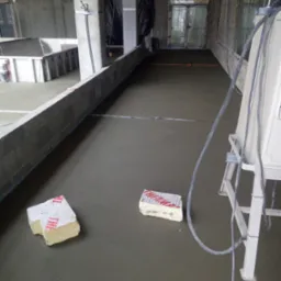 Wylewka posadzki w surowym wnętrzu, widoczne bloki styropianu i przewody maszyny wylewającej, betonowe ściany i słupy, okna w tle.