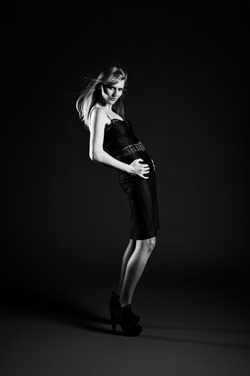 Czarno-białe zdjęcie modelki w sukience i butach na obcasie, pochylonej do tyłu, z rękami na biodrach, na ciemnym tle.