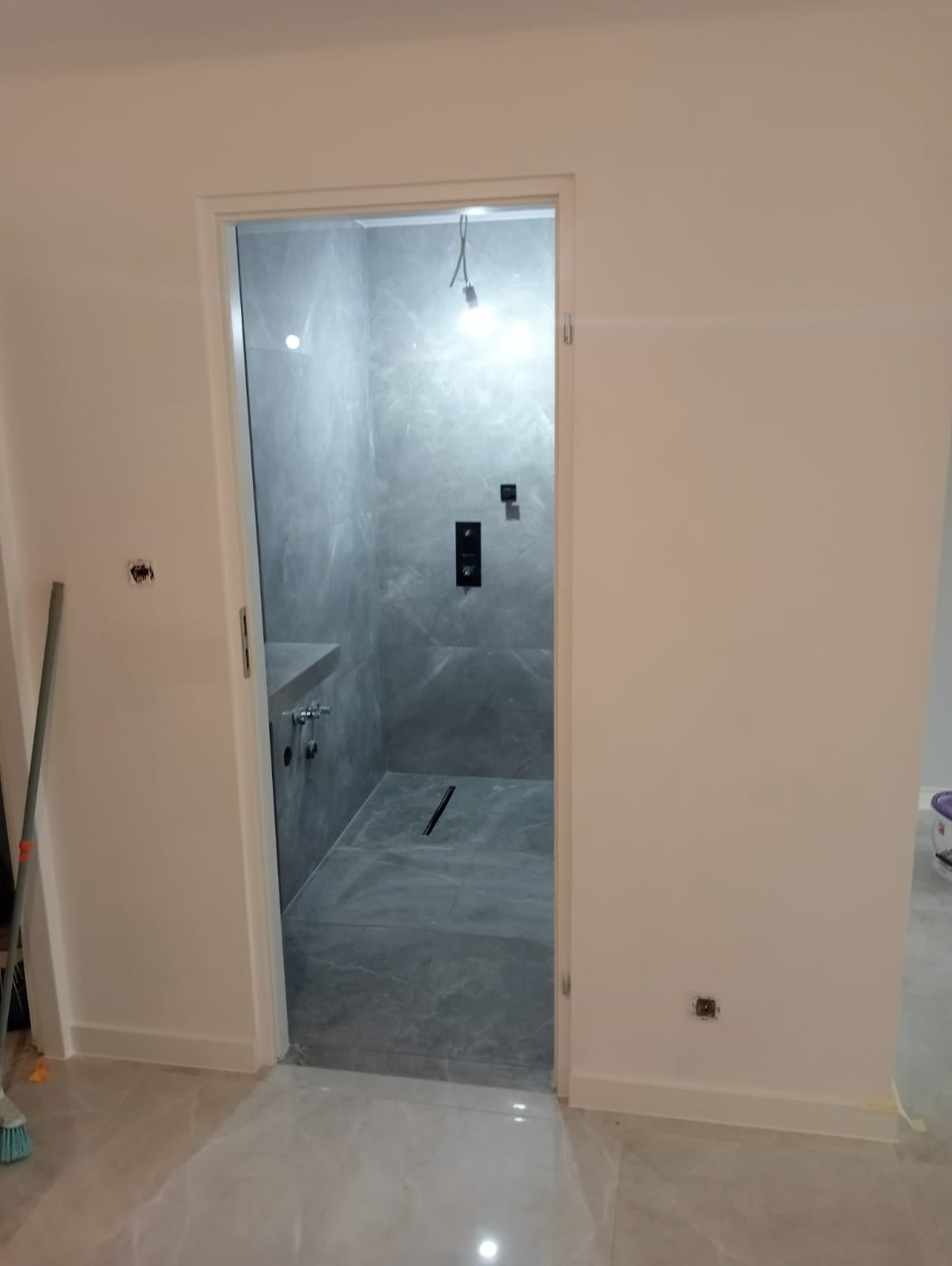 Nowoczesna łazienka z kabiną prysznicową typu walk-in wykończoną szarymi płytkami imitującymi marmur, widoczny odpływ liniowy i czarna armatura, surowe ściany w trakcie malowania.