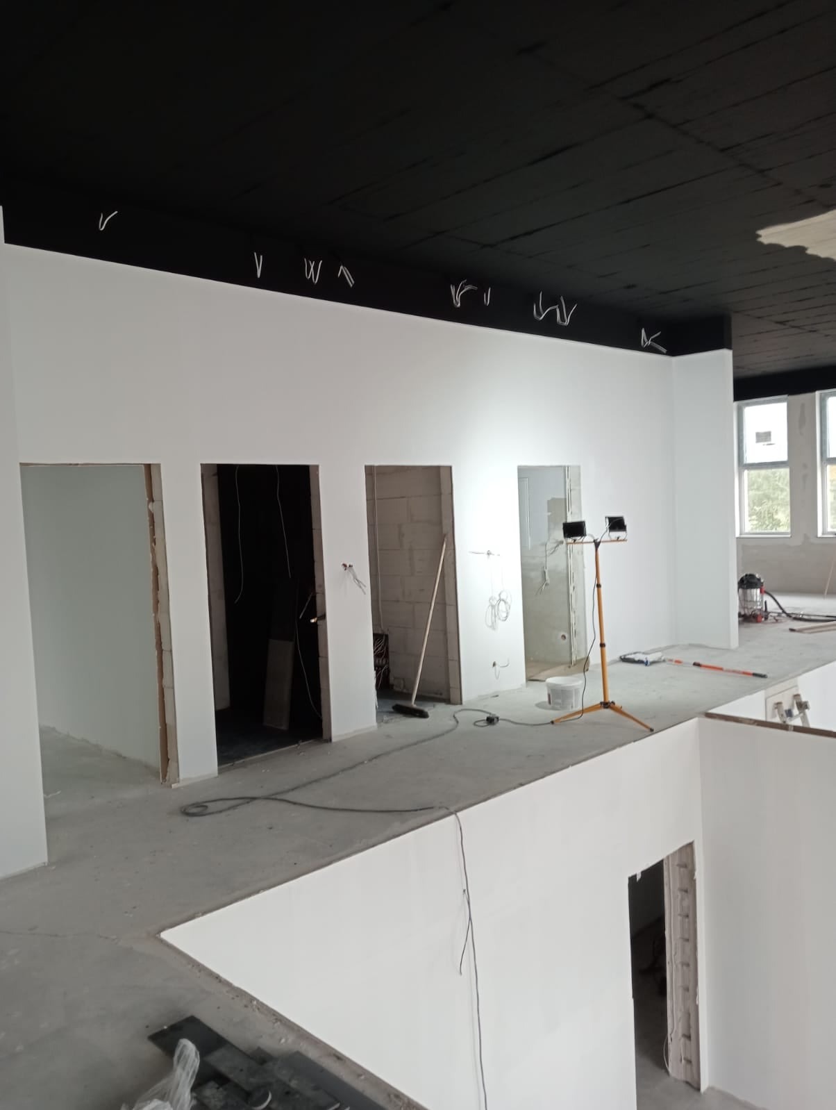 Wnętrze w trakcie malowania na biało, czarny sufit, widoczne kable, lampa robocza na statywie, podłoga betonowa, kilka otworów drzwiowych.