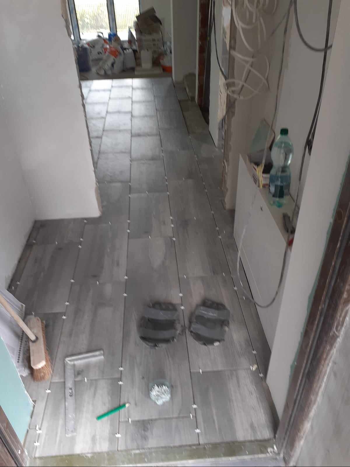 Układanie szarych płytek podłogowych imitujących drewno w korytarzu, widoczne krzyżyki dystansowe, narzędzia glazurnicze oraz dwa okrągłe wycięcia w płytkach.