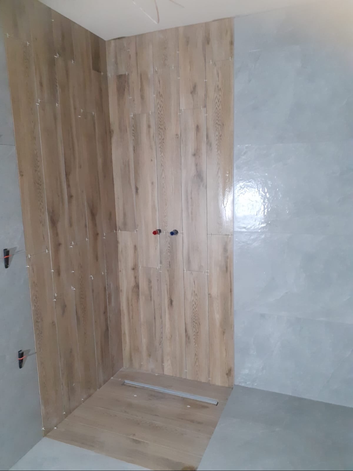 Ściany łazienki w trakcie układania płytek: imitacja drewna na jednej ścianie, szare płytki na drugiej, widoczne krzyżyki dystansowe i rurki doprowadzające wodę.