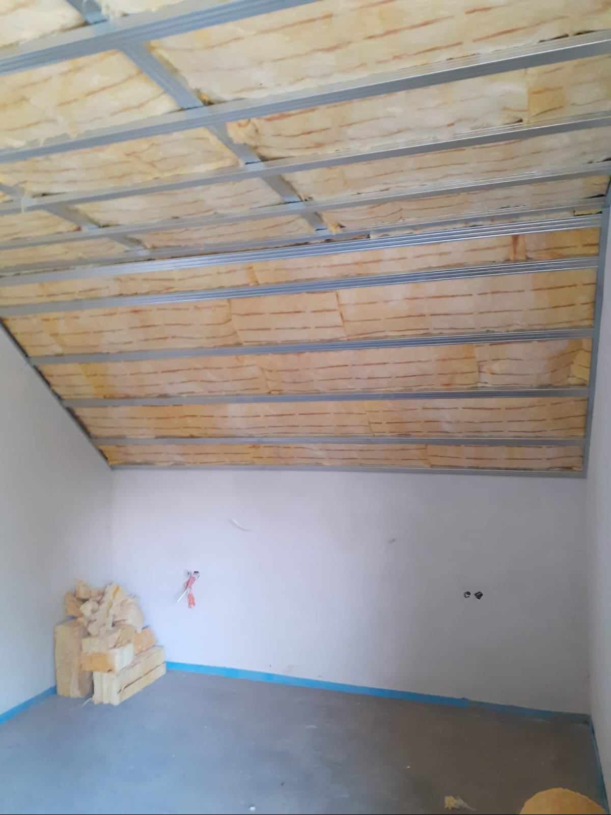 Montaż izolacji z wełny mineralnej w konstrukcji sufitu podwieszanego na poddaszu, widoczny stos materiału izolacyjnego przy ścianie, przygotowanego do dalszego montażu.