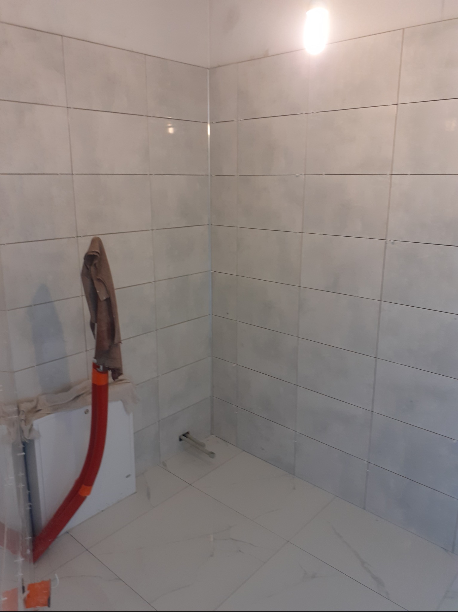 Świeżo wykafelkowana ściana i podłoga w łazience z widocznymi rurami instalacji wodnej i elementami zabudowy geberit, na ścianie robocza szmatka.