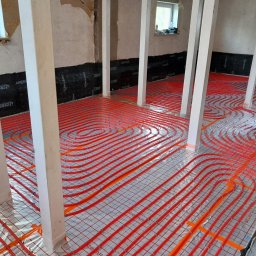 HYDRO-MANN - Czerwone rury ogrzewania podłogowego ułożone na folii izolacyjnej w pomieszczeniu z białymi filarami. Widoczne czarne maty izolacyjne na ścianach.